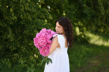 Dark-haired woman in a white dress standing sideways and looking at blooming pink peonies in a natural garden setting. Horizontal composition with soft daylight and a romantic spring atmosphere. © Anastasiia Harbar