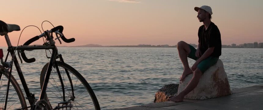 Static anamorphic wide shot of man in T shirt sitting on a stone by the sea and watching the sunset. Bicycle in foreground and calm water emphasize a thoughtful, peaceful coastal leisure mood.