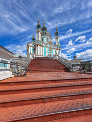 Naklejka premium Saint Andrew's Church in Kyiv under a bright blue sky with wispy clouds, featuring a grand red tiled staircase leading up to the historic baroque landmark.
