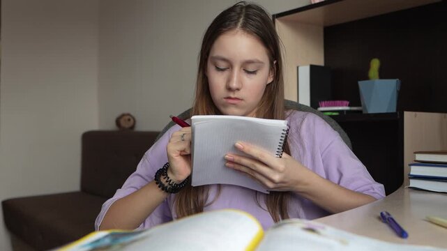 Schoolgirl copying text from textbook into notebook while preparing for lesson. Study process and homework preparation in home environment.