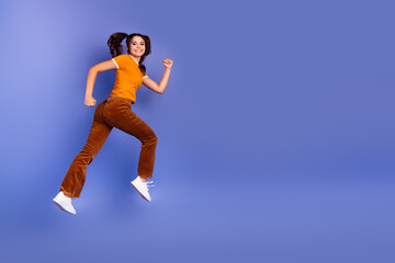 Naklejka premium Jumping young girl in orange shirt and brown pants against blue background expressing happiness and style