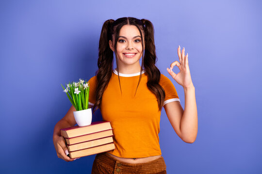 Young female student with book and plant smiling against purple background