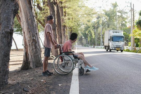 Volunteer or caregiver pushing young disabled man in wheelchair standing by public asphalt road near seaside, concept of outdoor mobility, health care support and community inclusion.
