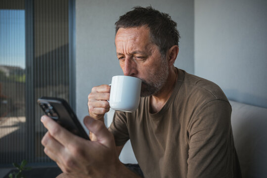 Middle-aged man drinking coffee while checking smartphone on balcony at home in morning light