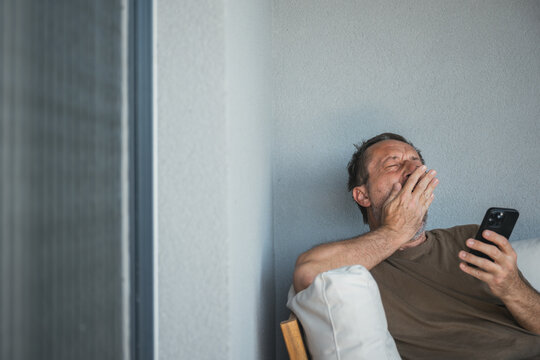 Tired middle-aged man sits on a balcony sofa, yawning and covering his mouth while checking his smartphone, conveying fatigue, boredom, late-night scrolling and everyday digital life.