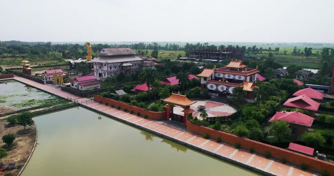 Aerial drone view of a Tibetan Buddhist monastery with an ornate gate and courtyard beside a still reflecting pond in the Lumbini Monastic Zone, Nepal.