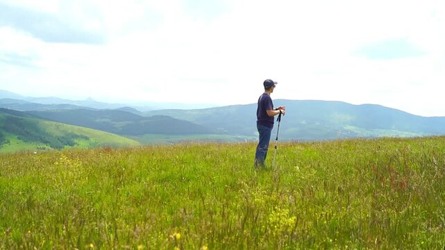 Hiker rests on mountain meadow holding trekking poles while observing layered ridges. Captures nature based relaxation, scenic retreat experience, and personal renewal through outdoor exploration.