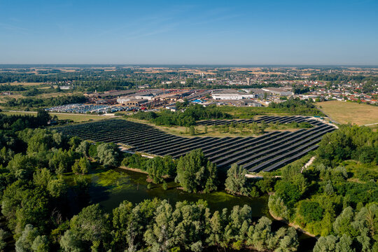 Aerial view of a solar farm with rows of photovoltaic panels near a pond and trees, adjacent to an industrial area in Saint-Denis-de-l'Hotel, Centre-Val de Loire, France.