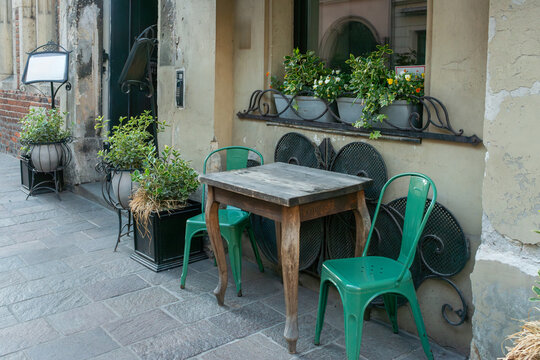 Rustic cafe terrace featuring weathered wooden furniture, green Tolix-style metal chairs, potted Euonymus fortunei shrubs, ivy and flowering window boxes against an aged historic facade.