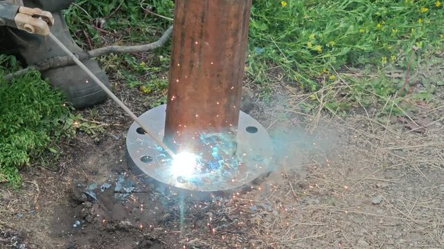 Close-up of professional welder welding steel pipe flange with electric arc creating bright sparks and smoke at construction site.