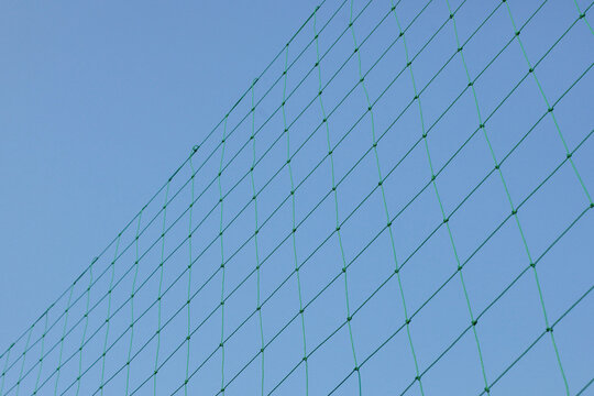 A low-angle shot looking up at a bright green diamond-patterned nylon mesh safety net stretched across a clear blue sky background.