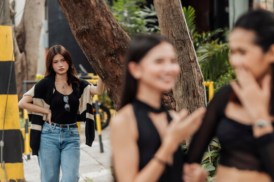 A jealous young Southeast Asian woman secretly watches her girlfriend with another woman at the park, showing suspicion, envy, and possessive behavior.