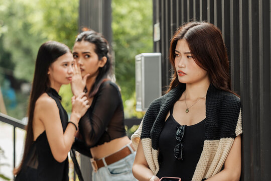 A young Southeast Asian woman looks uncomfortable as two women whisper behind her, capturing gossip, social tension, and feelings of exclusion