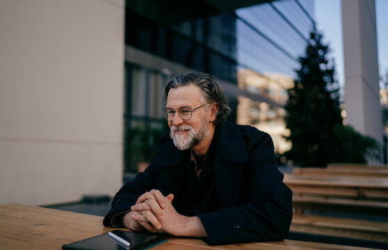 Successful mature professional sitting at outdoor cafe with digital tablet, confident middle aged businessman looking satisfied near modern architecture, financial freedom, career growth, ambition.