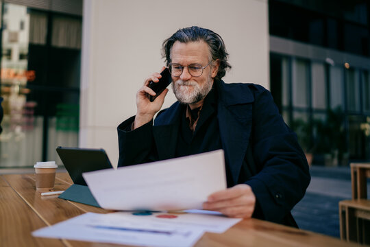 Senior male entrepreneur managing financial analytics on tablet and calling partner, mature professional discussing market trends while holding charts in street cafe, digital nomad remote work.