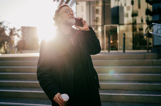 Ambitious senior man looking up with hope during phone call near modern office, mature professional in coat having successful mobile conversation in city under bright sunlight, copy space.