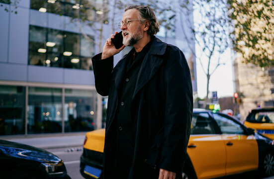 Smiling senior businessman talking on mobile phone standing in city street, happy mature male professional enjoying cellular conversation on smartphone near modern glass office buildings.