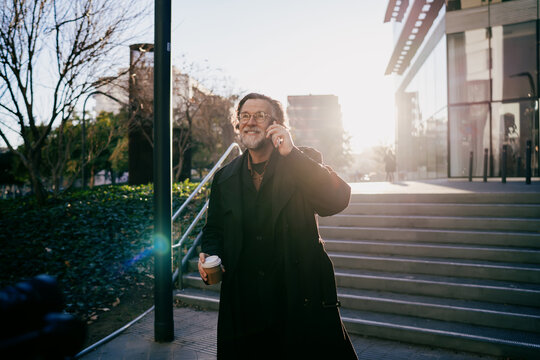 Cheerful senior entrepreneur walking in city park area with coffee having mobile phone call, mature digital nomad enjoying sunny morning and successful business communication, copy space.