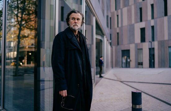 Serious senior businessman in black trench coat looking away while standing near glass office building, mature professional male manager thinking outdoors in modern urban business district.