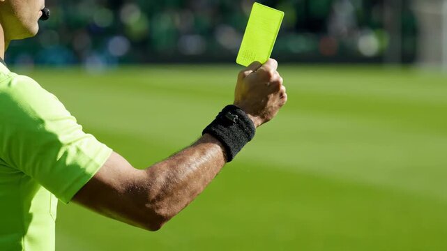 A referee raises a yellow card during a soccer match, signaling a penalty on a green pitch. The disciplinary gesture captures authority and tension in competitive football.