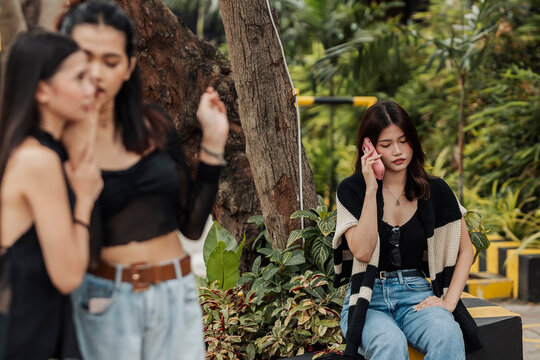 A young Southeast Asian woman sits alone talking on her phone unaware that two women talk behind her back and slander her.