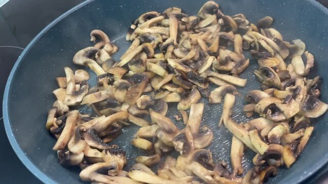 High angle horizontal view of sliced agaricus mushrooms frying in a modern nonstick pan on an induction stove, showing browning details and light steam during cooking. High quality 4k footage