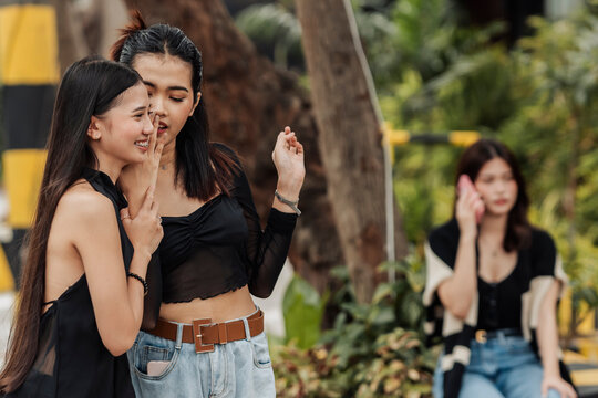 Two young Southeast Asian women whisper and share gossip while another woman sits alone using her phone, showing exclusion and social tension.