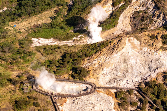 Aerial view of volcanic steam vents and geothermal activity with wooden boardwalks winding through lush green vegetation and rocky terrain in Indonesia.