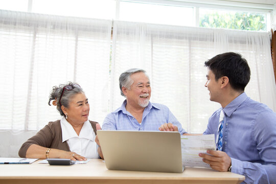 A cheerful elderly Asian couple sits at a desk, smiling while consulting a professional financial advisor or insurance agent holding documents in front of a laptop computer at home