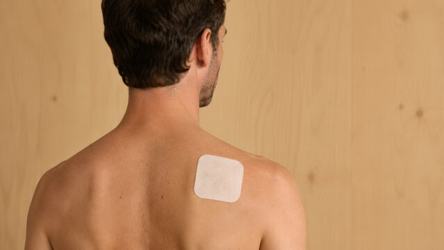 A man seen from the back, showing a square medical adhesive patch applied to his shoulder blade for treating muscle pain or injury in a professional setting