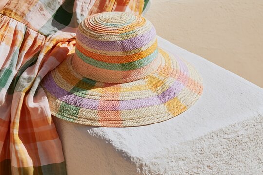 Close up of a woven straw sun hat with colorful stripes resting on a white surface next to a patterned dress in soft pastel colors