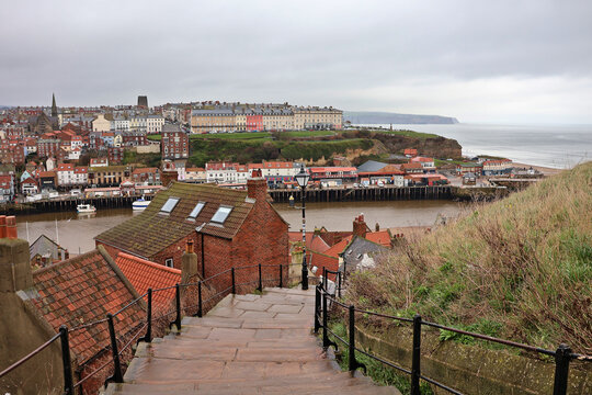 View along historic 199 Steps in Whitby with vintage street lamps, overlooking Whitby Harbour and Esk River, red rooftops of old town buildings on overcast day, North Yorkshire, England