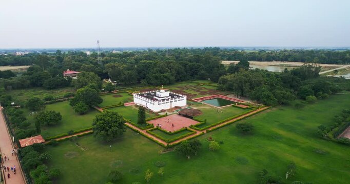 High aerial drone view of the white Maya Devi Temple surrounded by manicured gardens, sacred pond and vast green trees in Lumbini, Nepal.