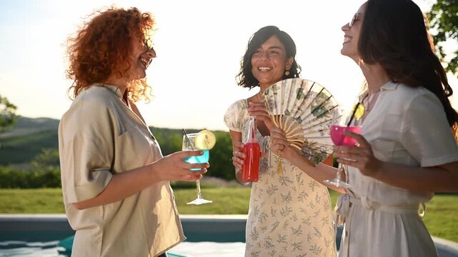 Three happy women laughing and toasting colorful cocktails together at a summer poolside party during sunset