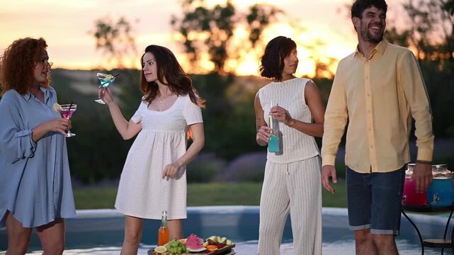 Group of friends dancing and enjoying cocktails during an outdoor sunset pool party with fruit and drinks on the table