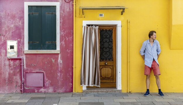 Young man in front of a house wall, red and yellow house facade with entrance door and window, colourful houses on the island of Burano, Venice, Veneto, Italy