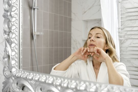 In a contemporary bathroom, a woman is focused on her skincare routine. Dressed in a soft robe, she examines her reflection in a decorative mirror while gently applying products to her face