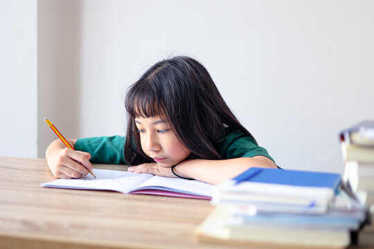 Focused young Asian girl studying at home, resting her arm on a wooden desk while diligently writing in a notebook with a pencil. Primary school student concentrating on homework.