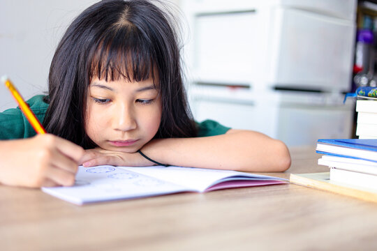 Focused young Asian girl studying at home, resting her arm on a wooden desk while diligently writing in a notebook with a pencil. Primary school student concentrating on homework.