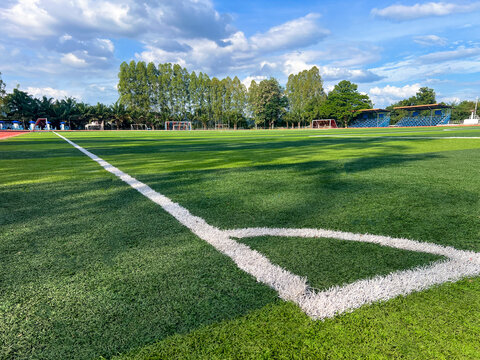 Wide angle view of green artificial grass soccer field with white corner line under a bright blue sky. Authentic outdoor stadium with stands and trees for sports competition and training backgrounds.