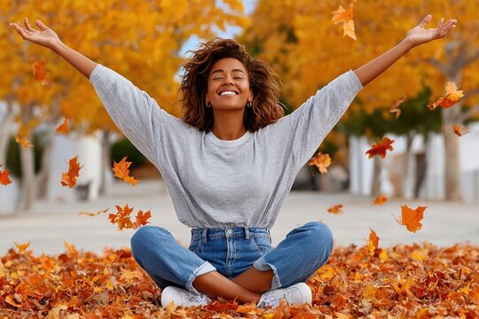 Smiling young woman sitting in autumn leaves with arms raised enjoying fall outdoors
