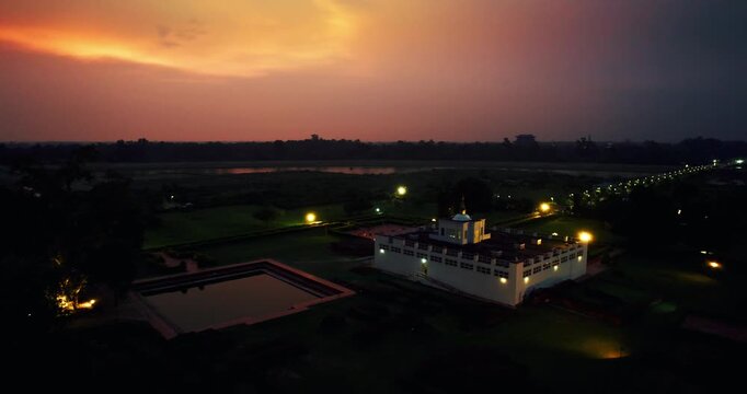 Aerial drone view of the illuminated Maya Devi Temple and its sacred pond in Lumbini, Nepal, under a glowing orange sunset sky at dusk.