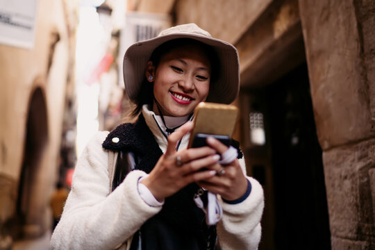 Happy Asian woman browsing social media feed on smartphone, smiling while reading news or chatting, 5G connection, urban lifestyle photography, authentic representation, focus on device.