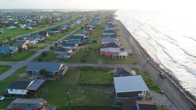 Seawall coastal frontage Surfside Beach corridor along Seashell Dr borders stone lined barrier with elevated homes facing Gulf horizon. Sunlit roofing, surf highlights extend in Freeport, Texas