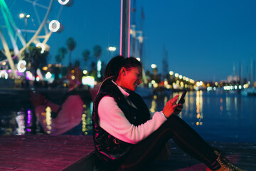 Obraz premium Asian woman using smartphone application on night city pier with neon lights. Smiling female tourist browsing social media on mobile phone with bokeh ferris wheel background, digital nomad lifestyle.