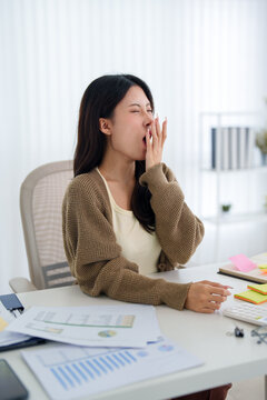 Young Asian businesswoman yawning with exhaustion during long work shift at bright modern office desk