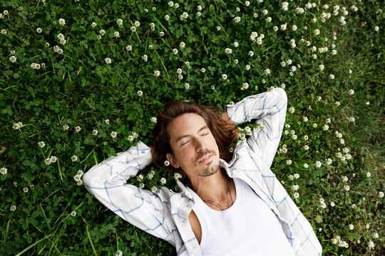 Upper view of handsome brutal hipster man lying on grass among blooming flowers in plaid shirt with closed eyes, having nap or daydreaming putting hands under his head, relaxing and enjoying peace