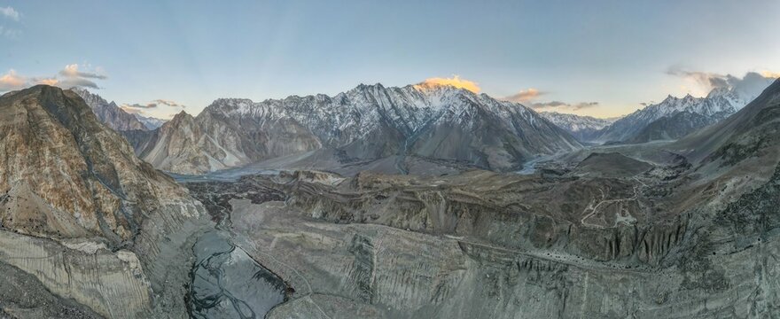 Aerial view of the rugged Karakoram Range with snow-capped peaks, deep valleys, and winding riverbeds under a soft sunset sky in Hunza Nagar, Gilgit, Pakistan.