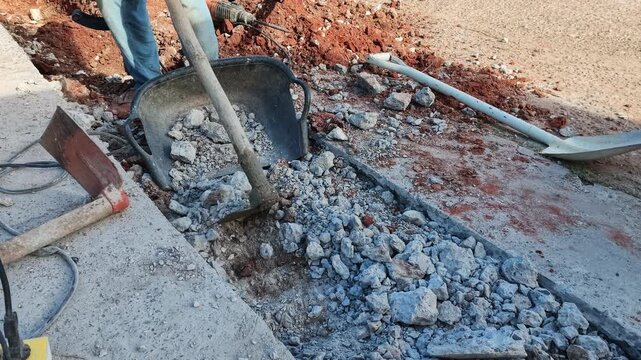 A close-up photograph of a worker using a hoe to shovel broken concrete and rocks into a black construction basket at a job site