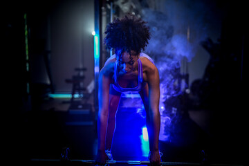 Obraz premium A woman in purple sportswear preparing for a heavy lift with a barbell in a moody, low-light gym environment with dramatic highlights.